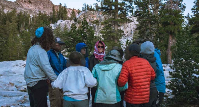 A group of people stand in a circle with linked arms. There are large rock formations and evergreen trees nearby. 